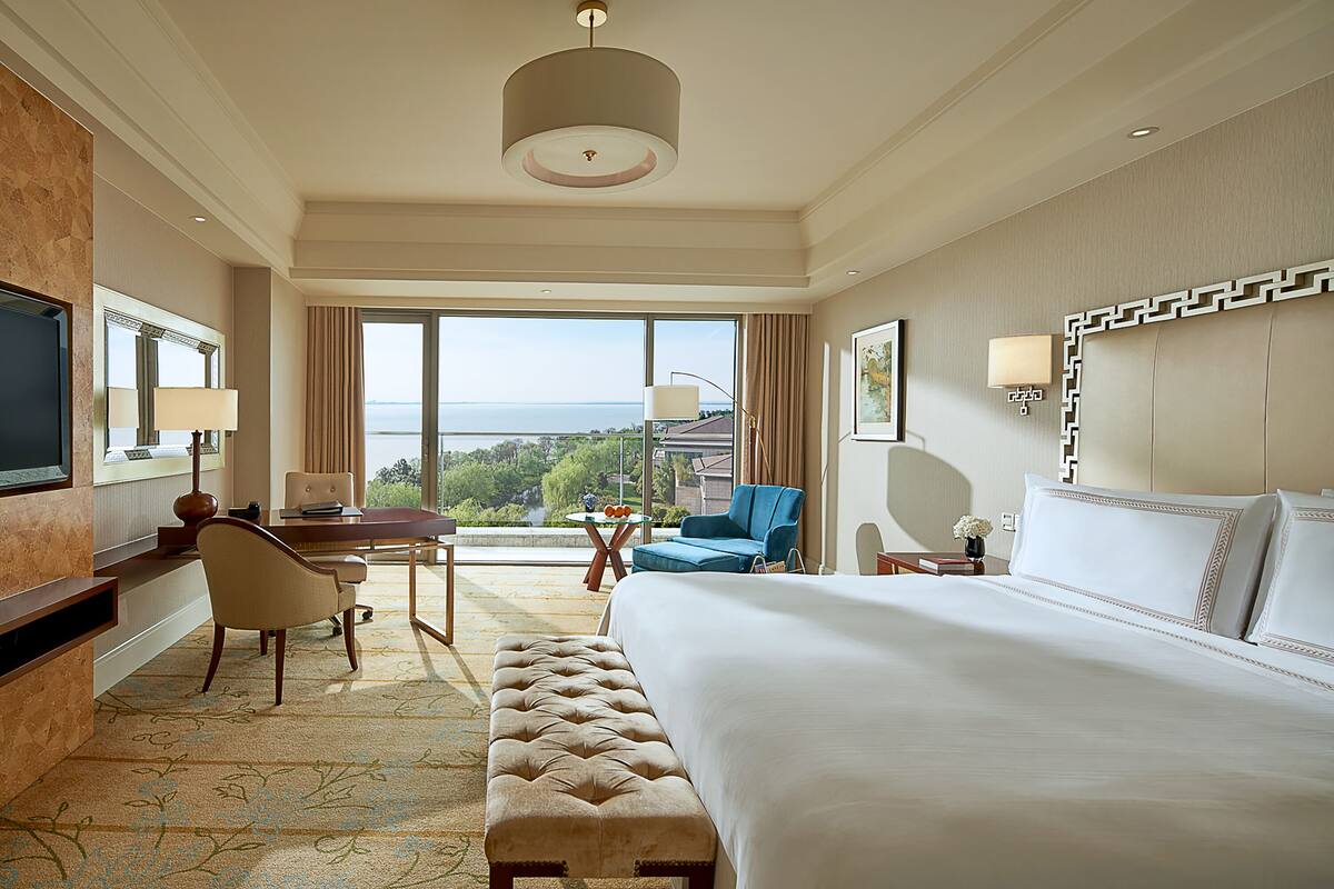 A hotel room with a large bed, a desk with a chair, a blue armchair, and a view of the sea.