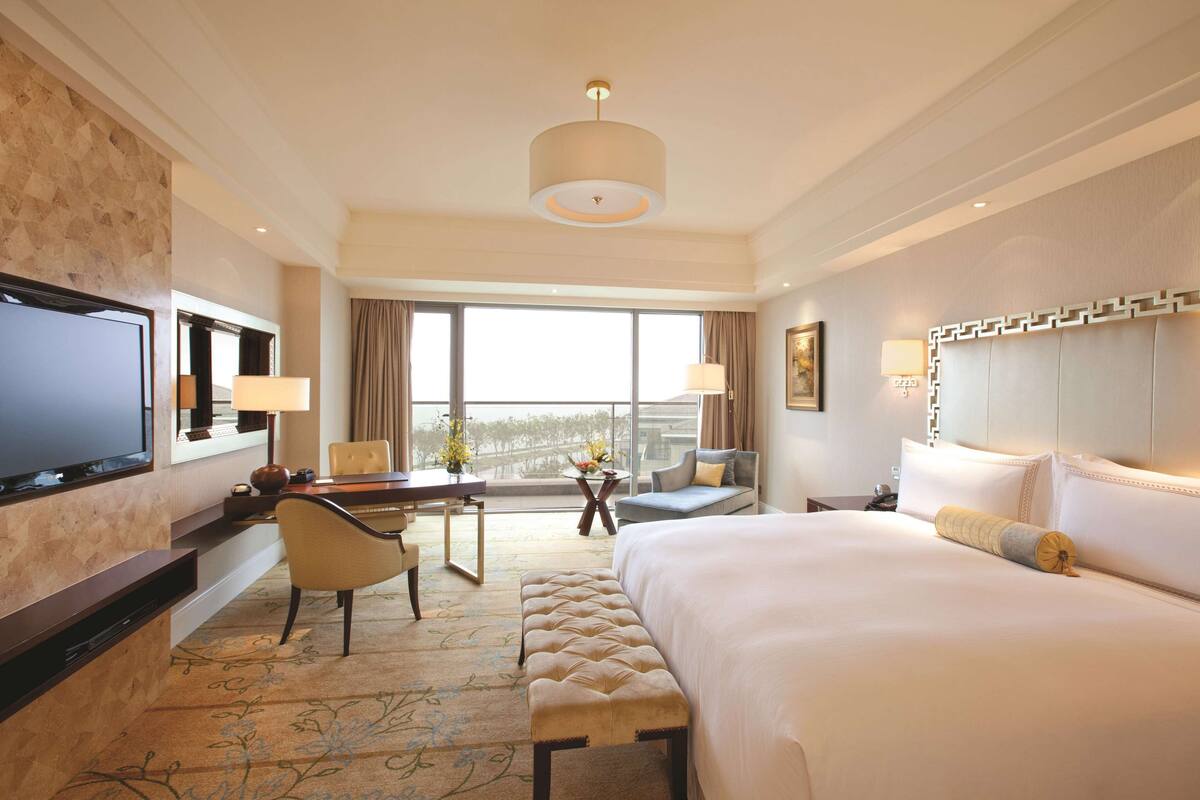 A hotel room with a large bed, a desk with a chair, a television, and a view of the outdoors through large windows.