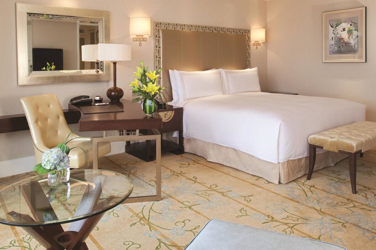 A hotel room with a bed, a desk with a lamp, a chair, a small table, and a bench.