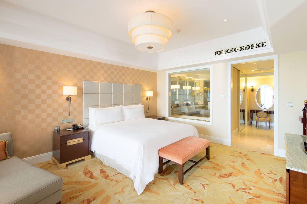 A hotel room with a large bed, bedside tables, a bench, a mirror, and a bathroom visible through an open door.