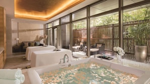 A spa room with a large bathtub filled with water and rose petals, a sofa with cushions, a small table with a tray, and a view of greenery through large windows. - Fairmont Yangcheng Lake (Suzhou)