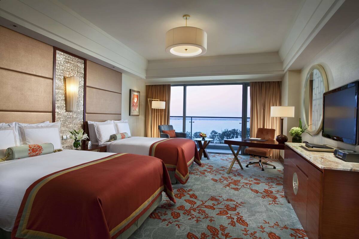A hotel room with two beds, a desk, a television, and a large window offering a view of the sea.