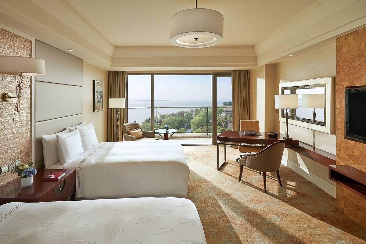 A hotel room with two beds, a desk, a chair, a TV, and a large window offering a view of the sea.