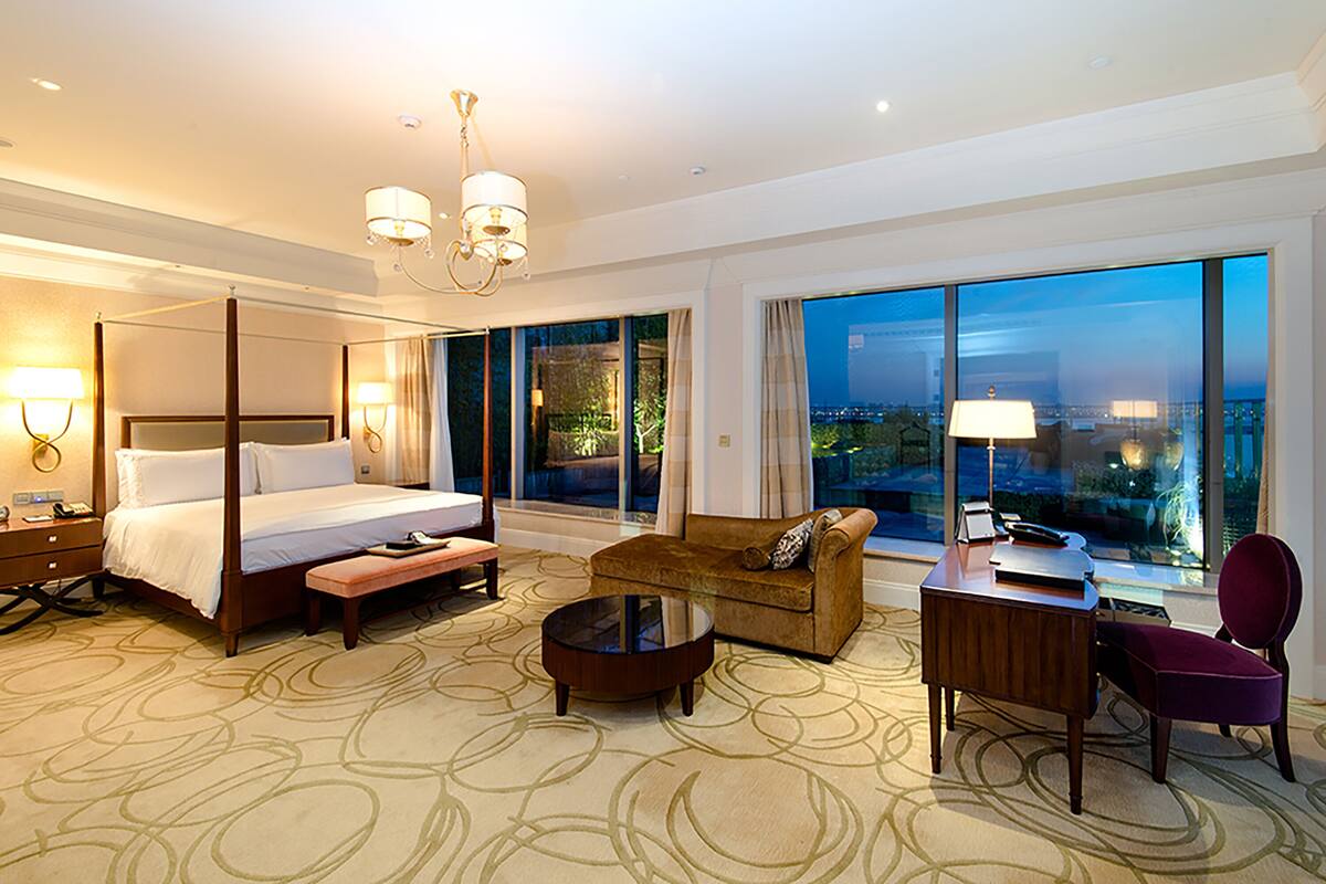 A hotel room with a four-poster bed, a sofa, a desk, and a chair. There is a large window with a view of the city.