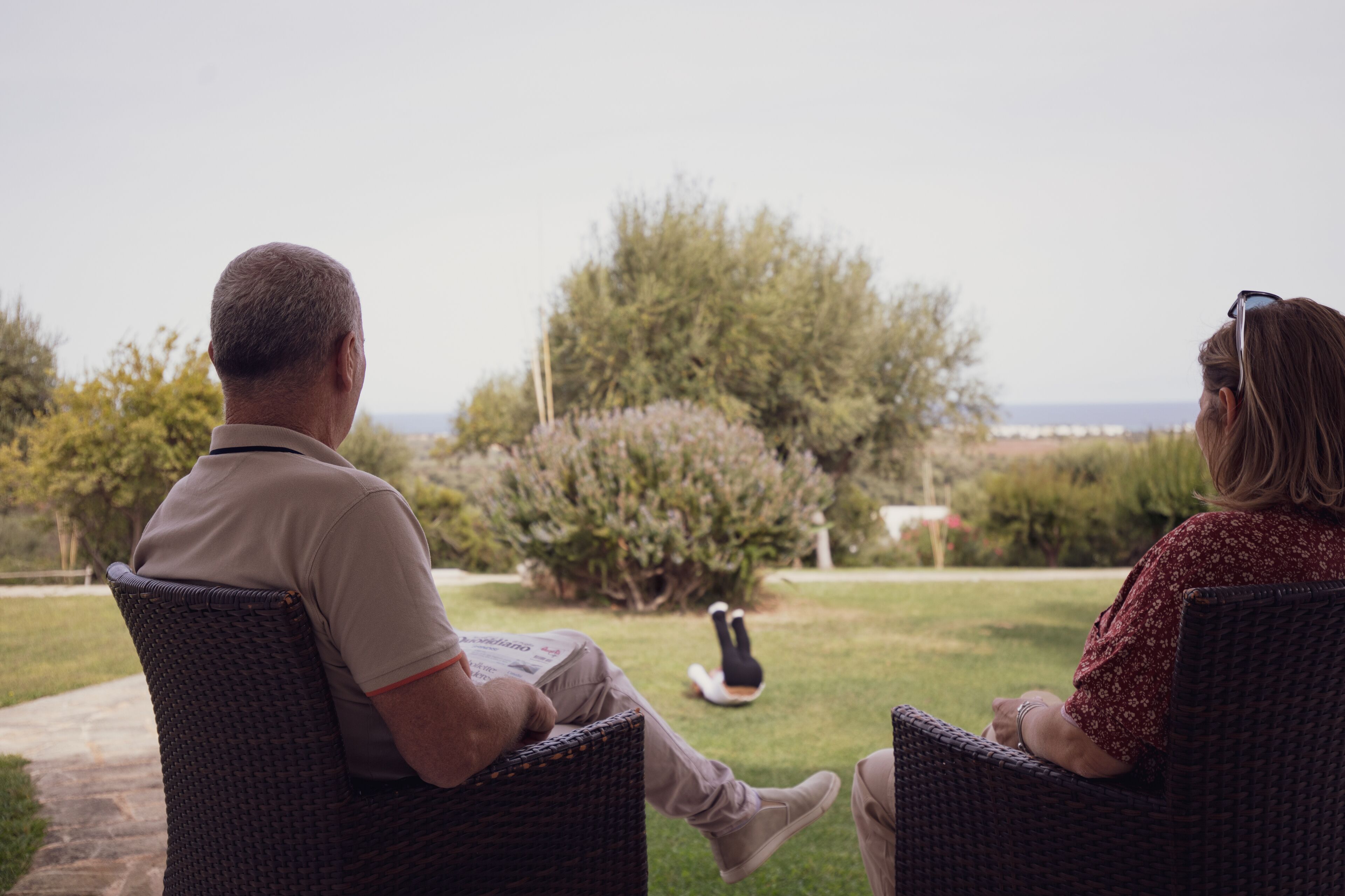 Dos personas sentadas en sillas de mimbre mirando un jardín con árboles y vistas al mar.