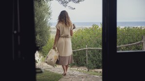 A woman in a beige dress walking on a stone path towards a beach, carrying a bag and a book. - Hotel Resort Corte Di Ferro (Carovigno)