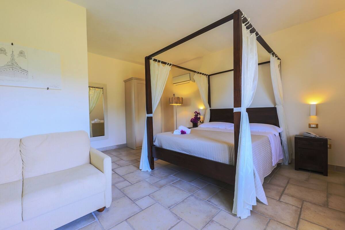A four-poster bed with white linens, a bedside table with a lamp, and a sofa in a well-lit room with tiled flooring.