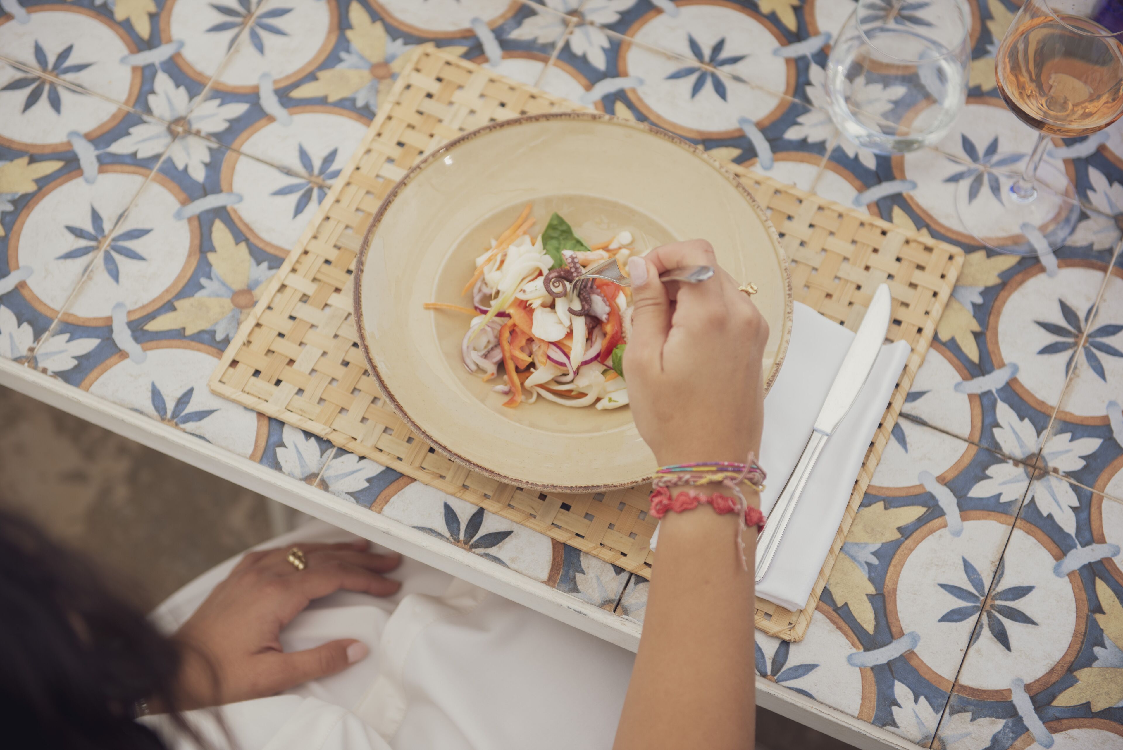 A table with a patterned tablecloth, a woven placemat, a plate of food, and a glass of wine.