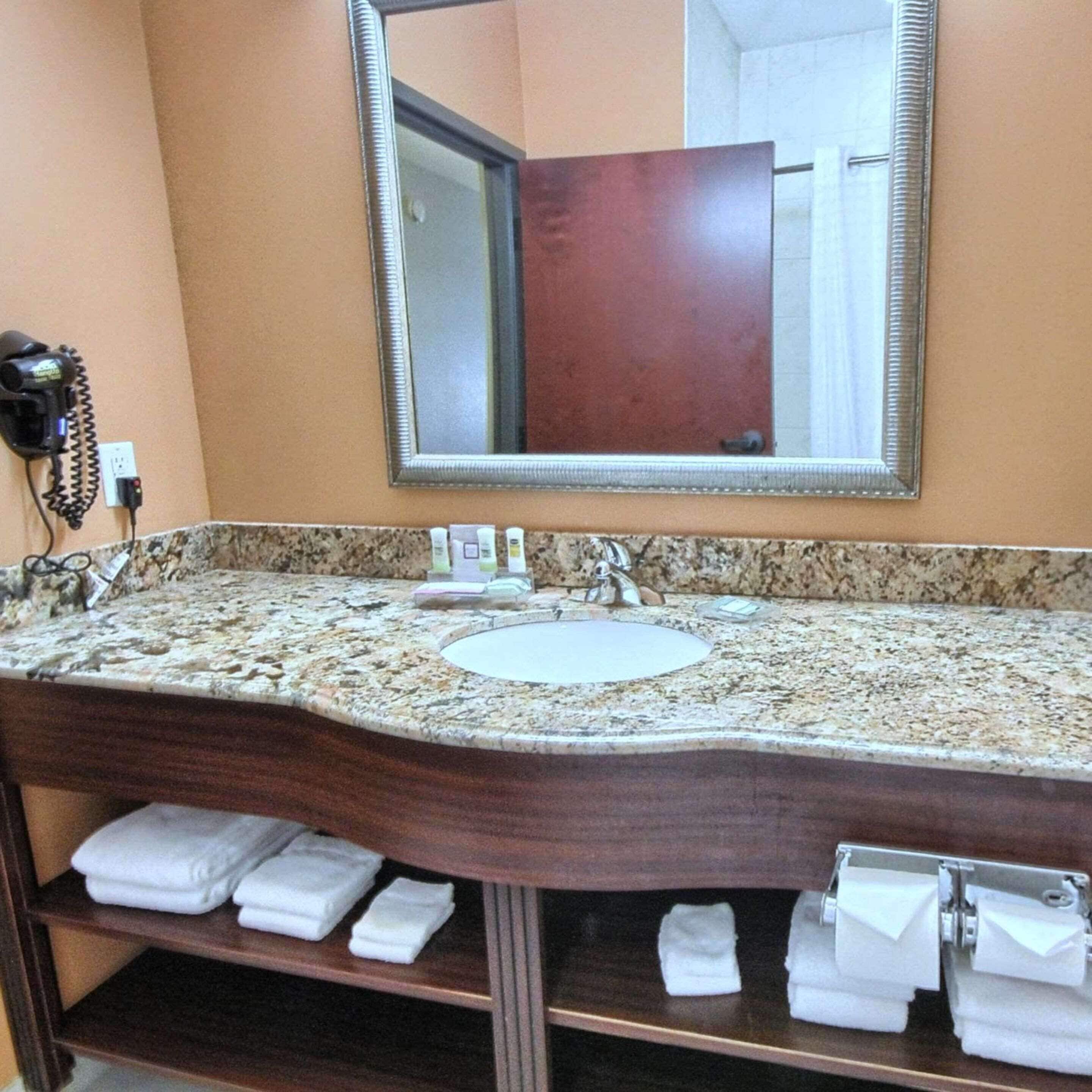 A bathroom with a granite countertop sink, a large mirror, and a hairdryer on the wall. There are towels on a shelf below the sink.