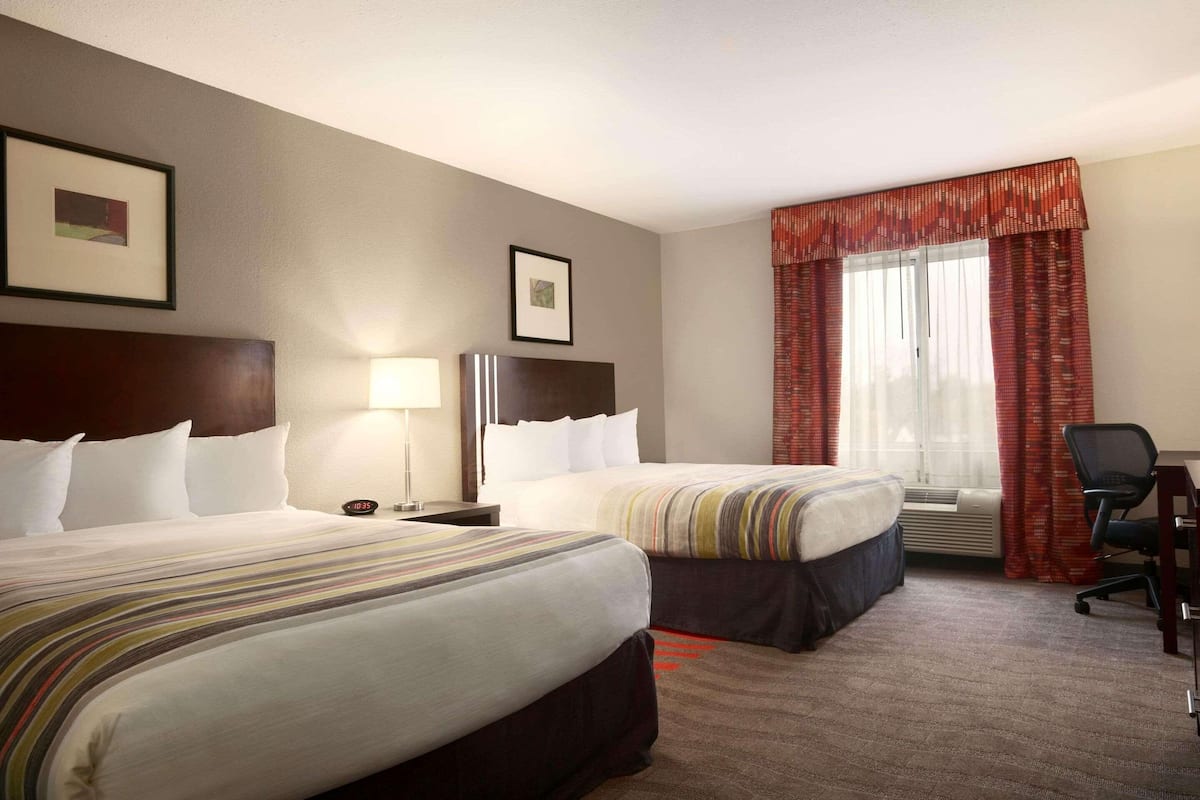 Une chambre d’hôtel avec deux lits, un bureau, une chaise, une lampe et une fenêtre avec des rideaux.