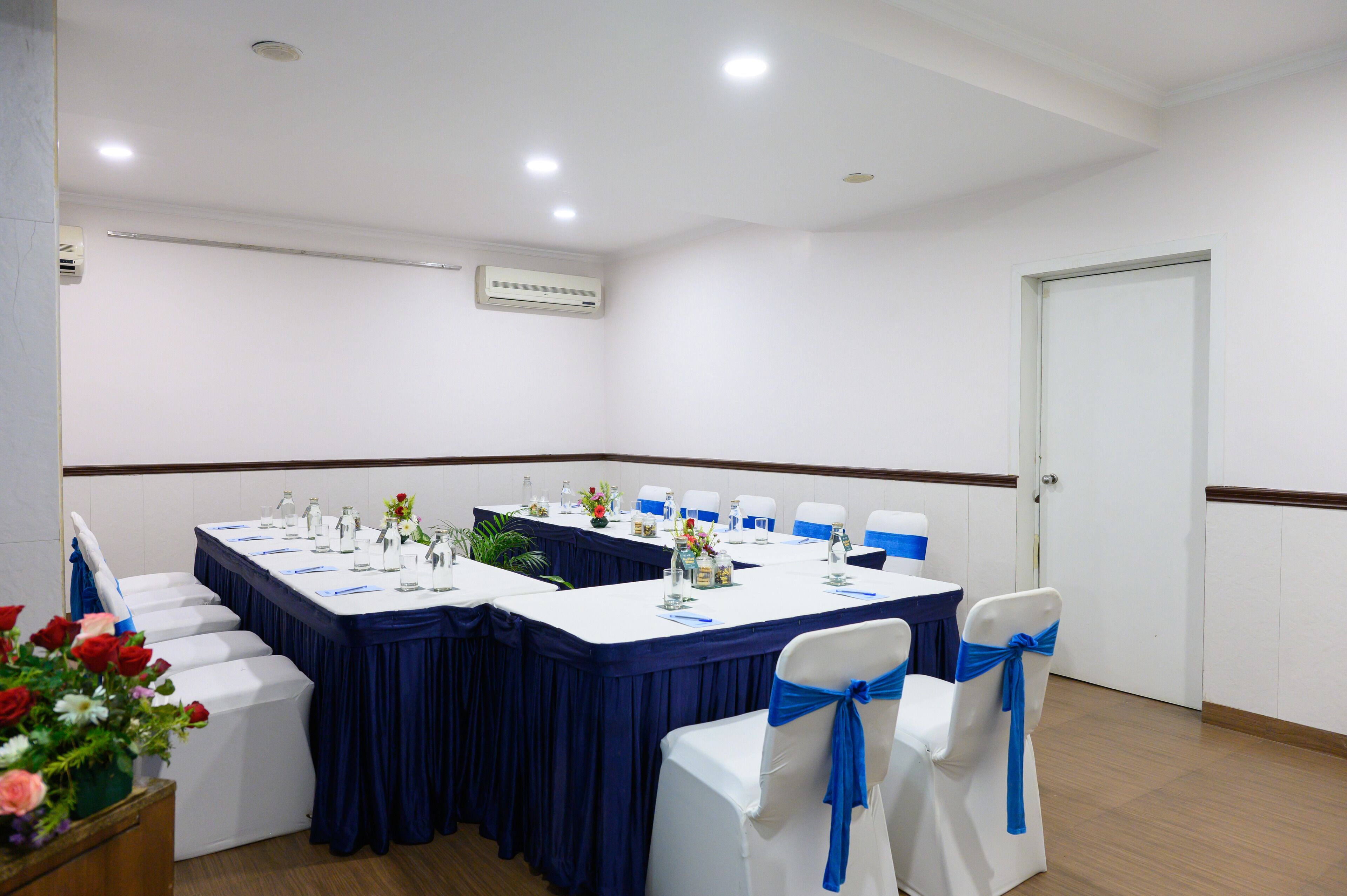 A conference room with a long table, white chairs with blue sashes, and a white door.
