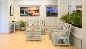 A well-lit room with white paneled walls, two framed landscape paintings, wicker chairs with patterned cushions, a glass coffee table, and a small side table with a lamp. - Sinclairs Siliguri (Siliguri)