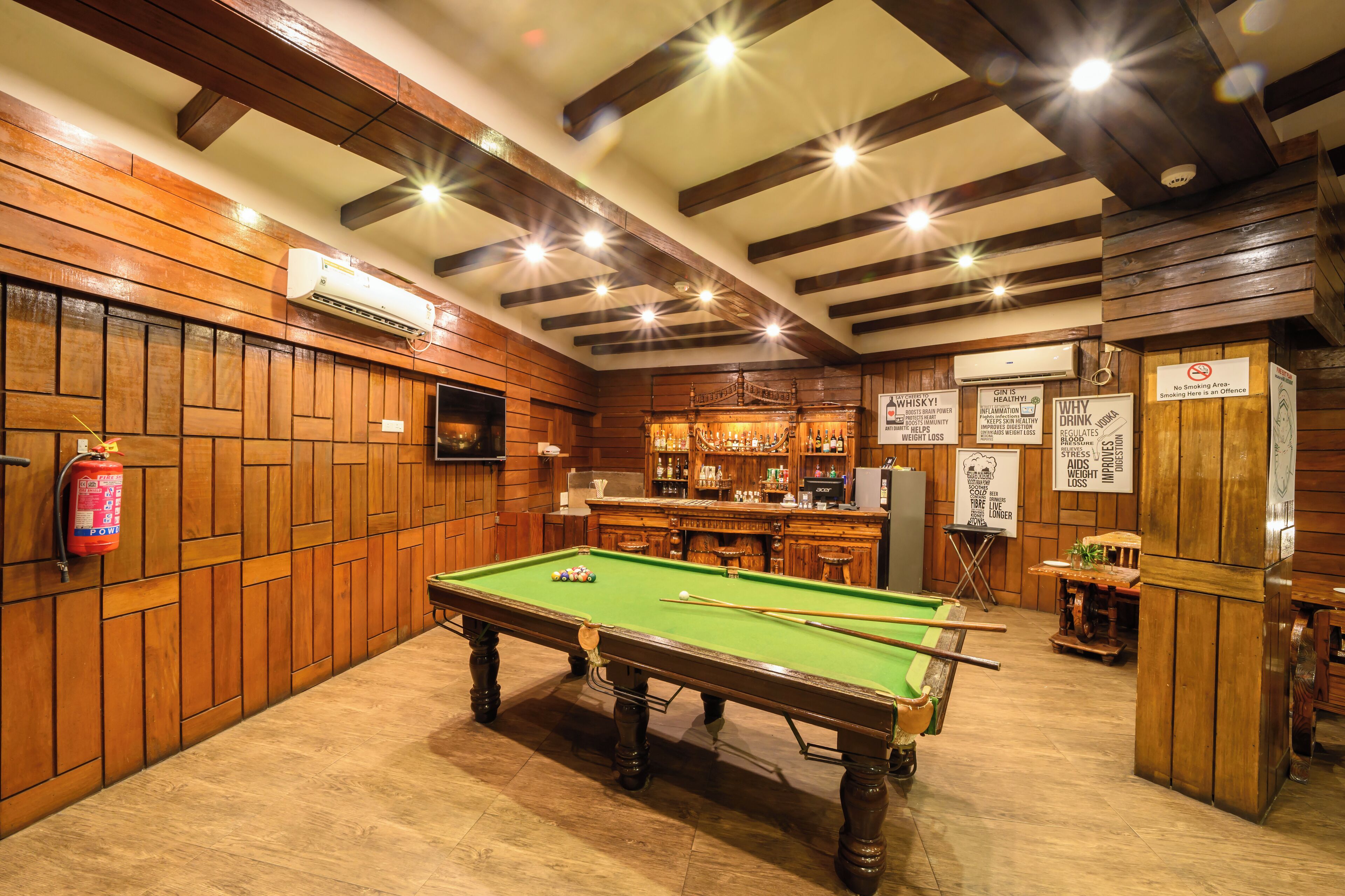 A pool table in a room with wooden walls and ceiling beams, a bar area with various bottles, and a fire extinguisher on the wall.