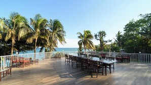Um deck externo com piso de madeira, rodeado por palmeiras e corrimão branco, com vista para um corpo de água.