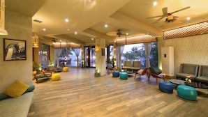 Um hall amplo com piso de madeira, várias áreas de estar incluindo sofás e cadeiras, ventiladores de teto e janelas grandes que permitem entrada de luz natural.