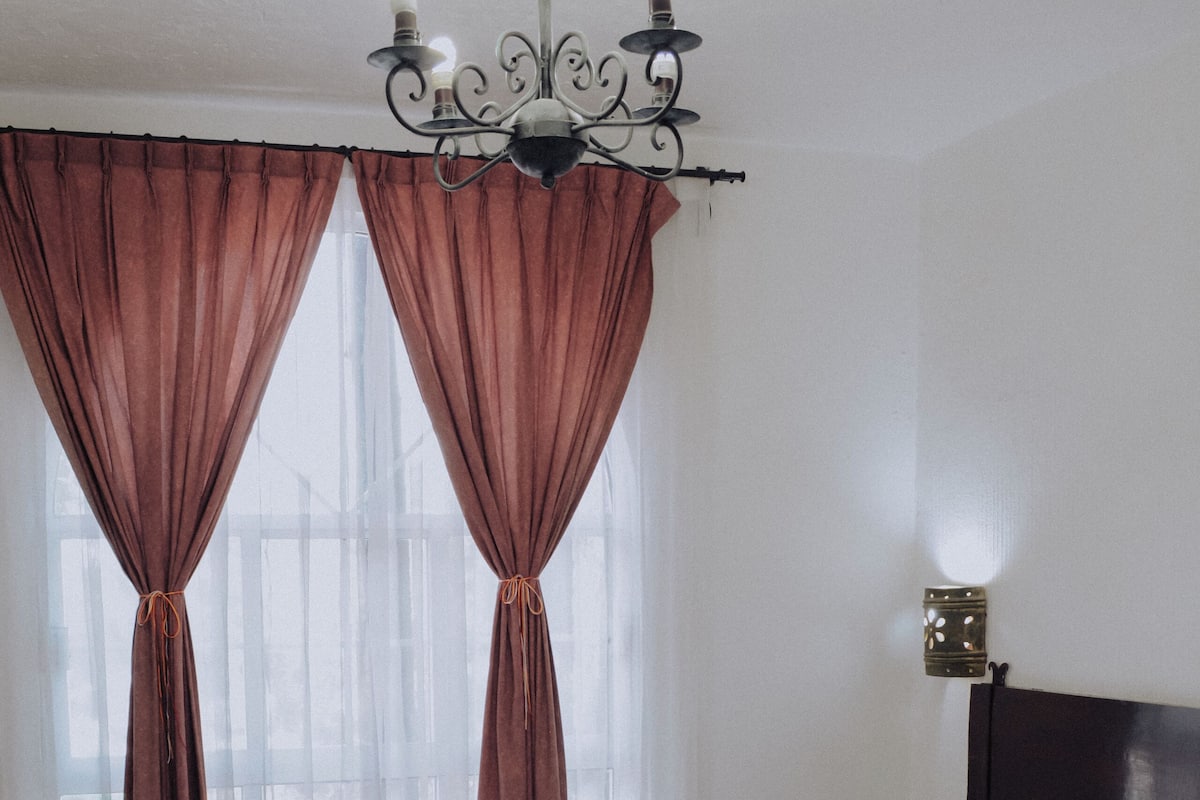A hotel room with a bed, curtains, a chandelier, and a nightstand.