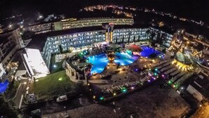 Ein großes, beleuchtetes Hotel mit einem zentralen Poolbereich, umgeben von einem angelegten Garten und einem Parkplatz.