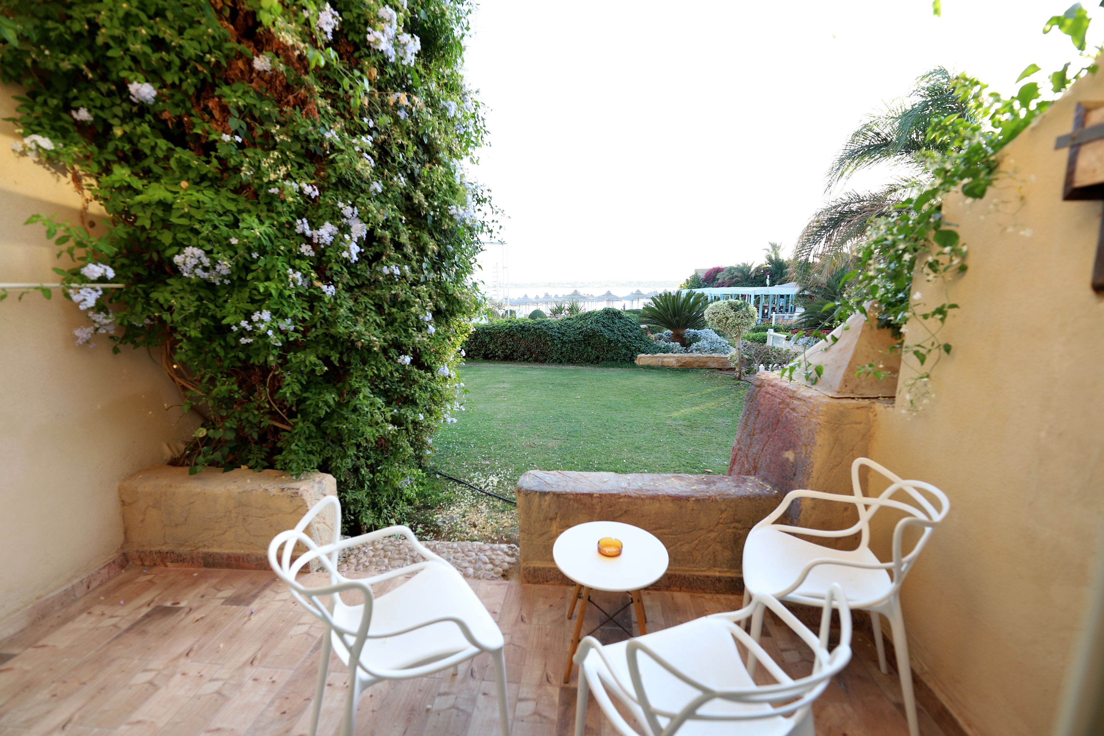 A patio with white chairs and a small table, surrounded by lush greenery and a view of a grassy area.