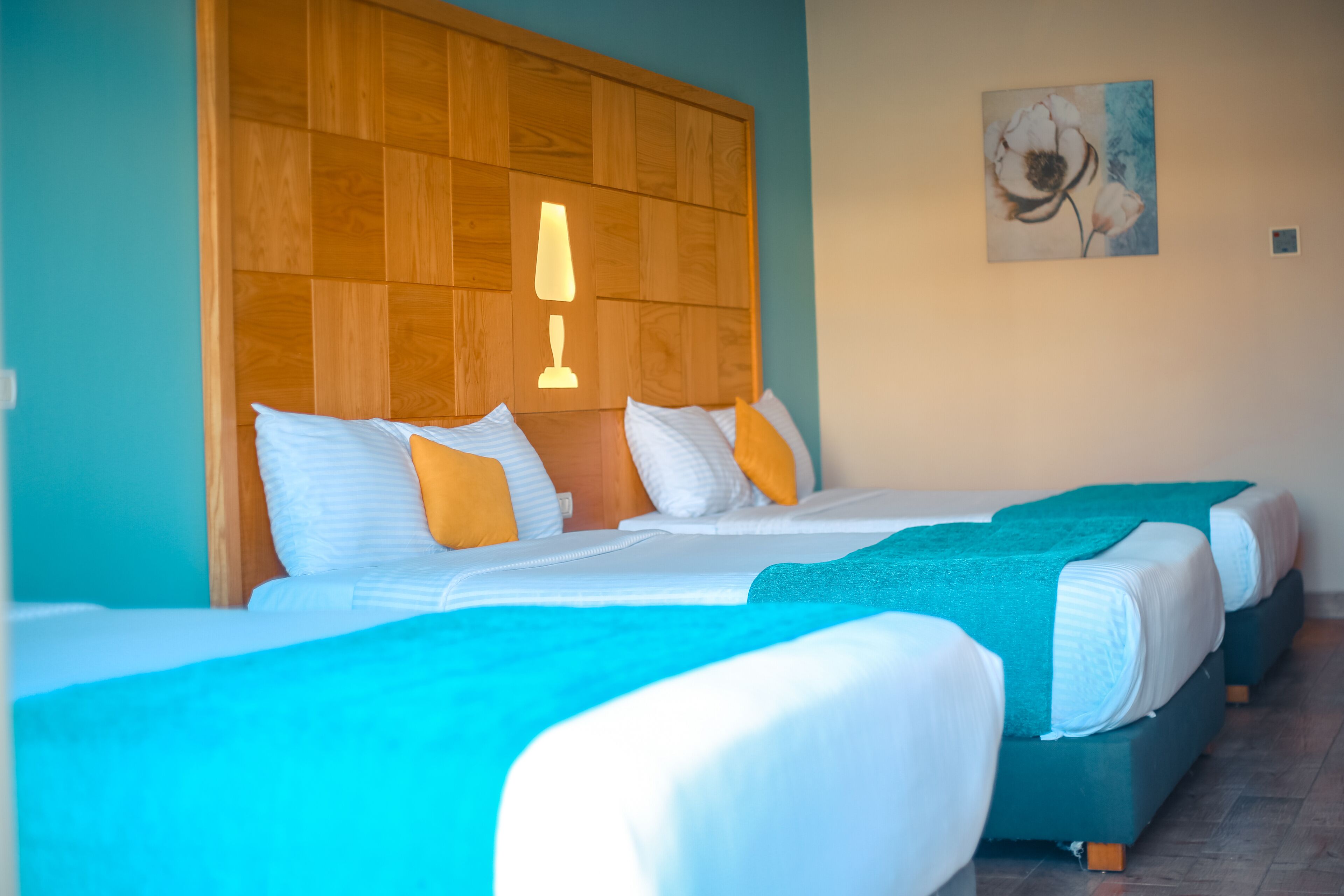 Une chambre d’hôtel avec trois lits, une tête de lit en bois, une lampe fixée au mur et un tableau.