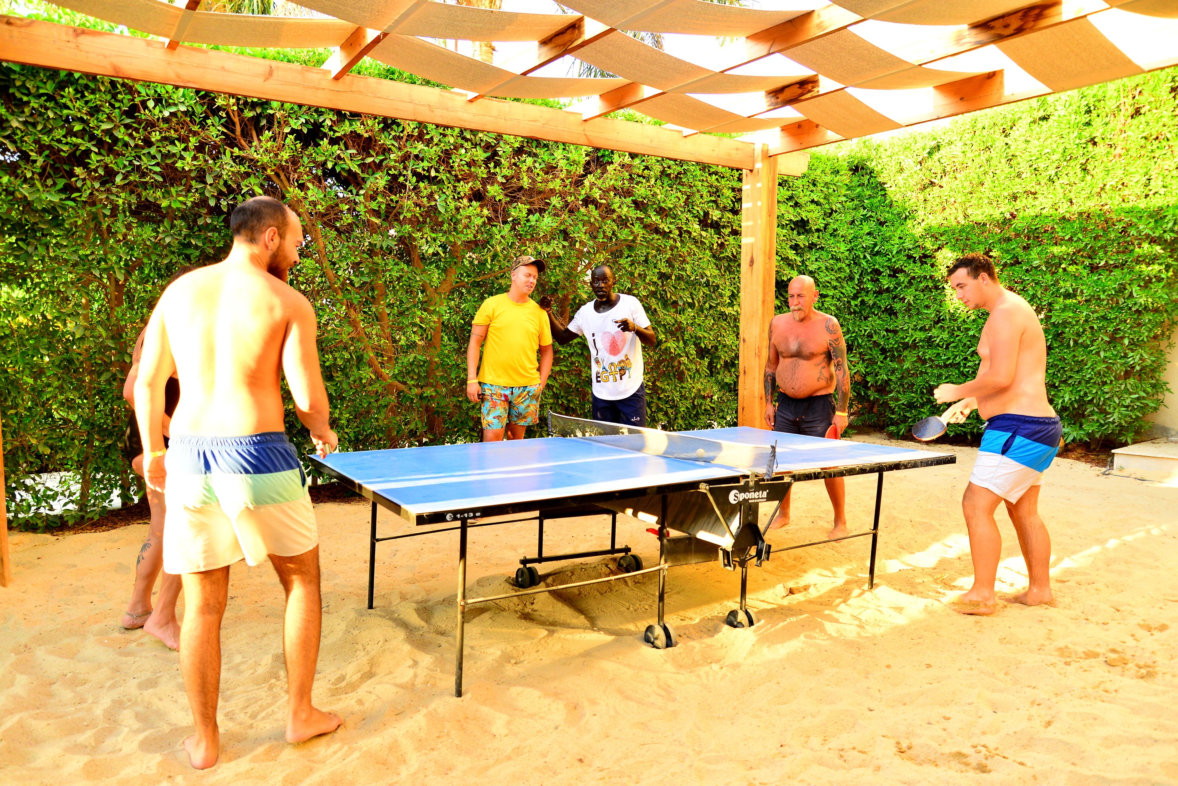 Un grupo de personas jugando tenis de mesa al aire libre sobre arena, bajo una pérgola de madera.