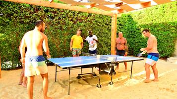 Un groupe de personnes joue au ping-pong à l’extérieur, sur une surface sablonneuse, sous une pergola en bois.
