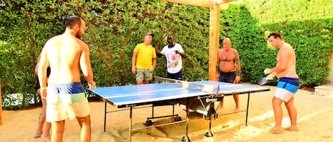 Un grupo de personas jugando tenis de mesa al aire libre sobre arena, bajo una pérgola de madera.