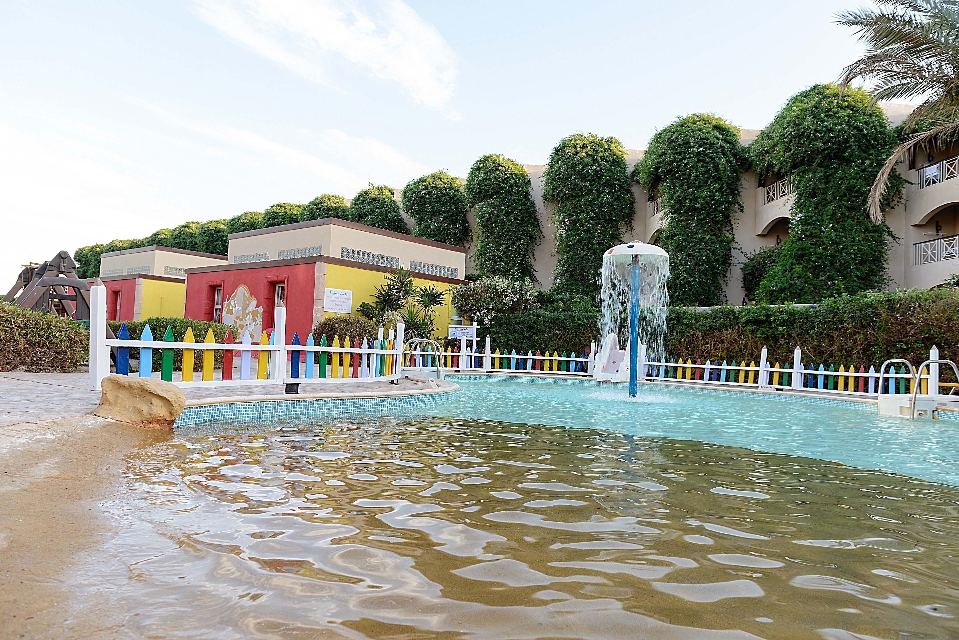 A swimming pool with a fountain, surrounded by a white fence with colorful posts, and buildings with greenery on the walls.