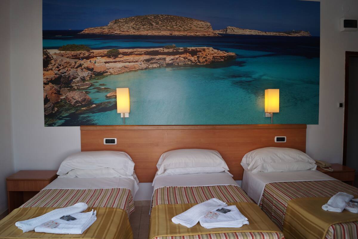 A hotel room with three beds, each with a striped bedspread and white pillows. There are two wall-mounted lamps on either side of the beds.