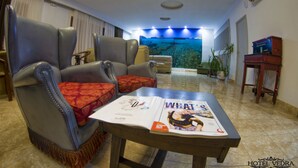 Habitación de hotel con zona de estar que incluye dos sillones de cuero con cojines rojos, una mesa de centro de madera con una revista y un gran acuario en la pared.