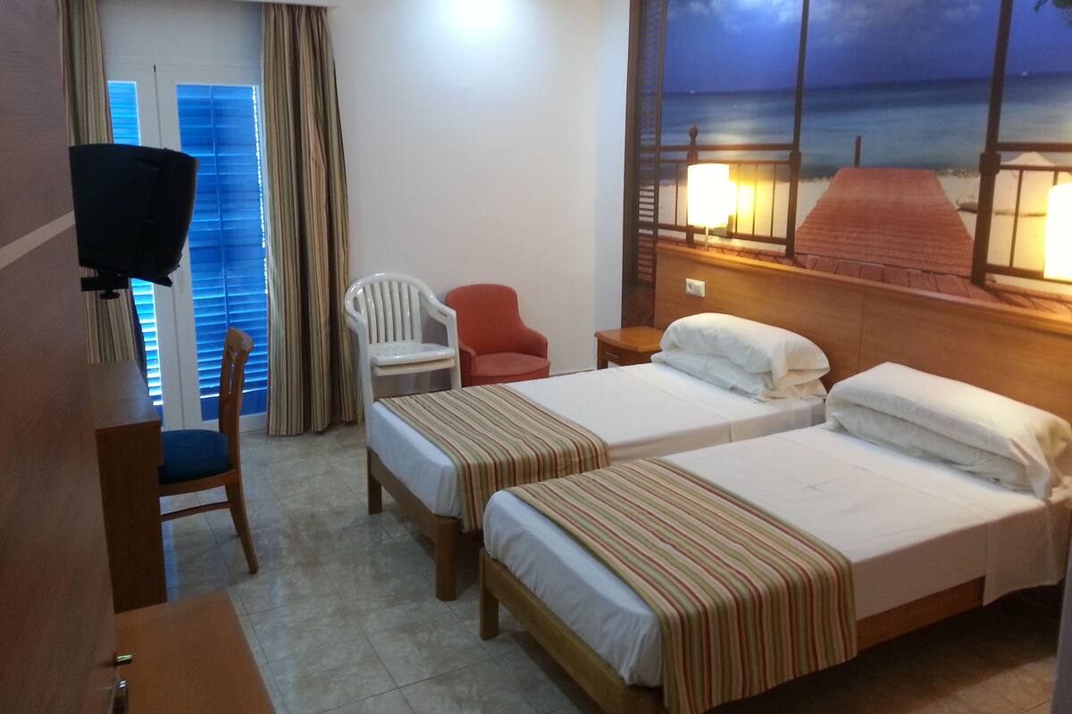 A hotel room with two beds, a TV, a desk, a chair, and a view of the sea at night.
