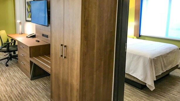Une chambre d’hôtel avec une armoire en bois, un bureau avec une chaise, un lit recouvert d’une courtepointe blanche, un téléviseur fixé au mur et une fenêtre avec des stores.