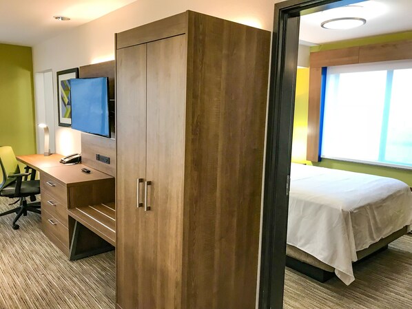 A hotel room with a wooden wardrobe, a desk with a chair, a bed with a white bedspread, a television on the wall, and a window with blinds.
