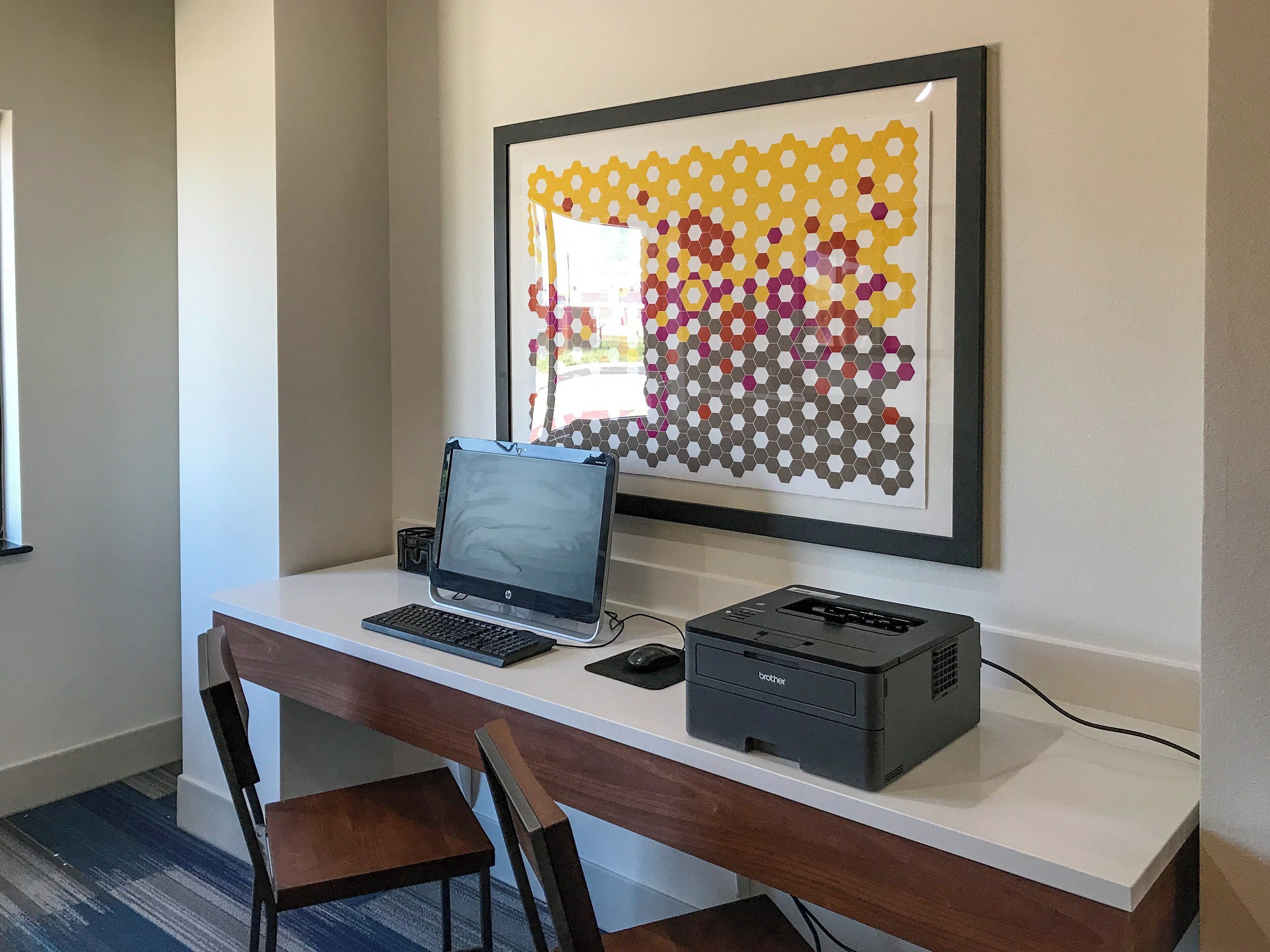 A workspace with a laptop, desktop computer, printer, and a framed abstract art piece on the wall.