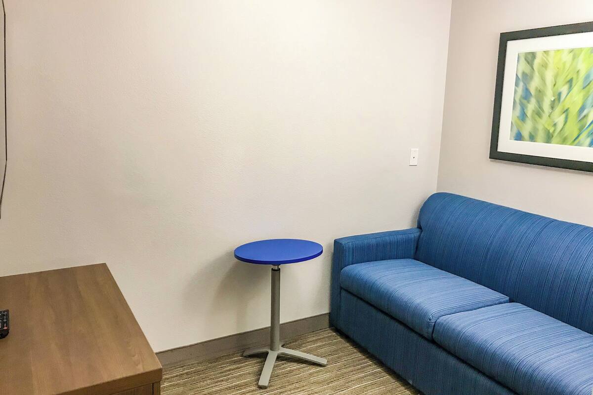 A room with a blue sofa, a small round table, and a remote control on a wooden side table.