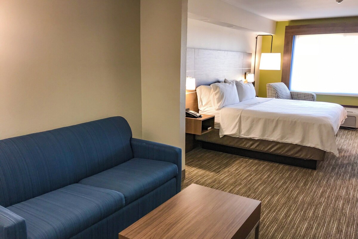 A hotel room with a blue sofa, a wooden table, a bed with white bedding, and a nightstand with a lamp.