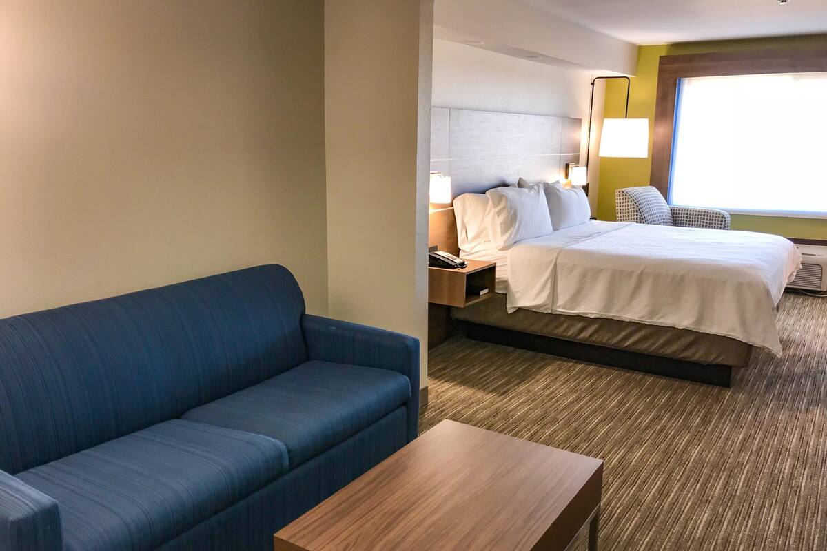 A hotel room with a blue sofa, a wooden table, a bed with white bedding, a nightstand with a lamp, and a window with a view.