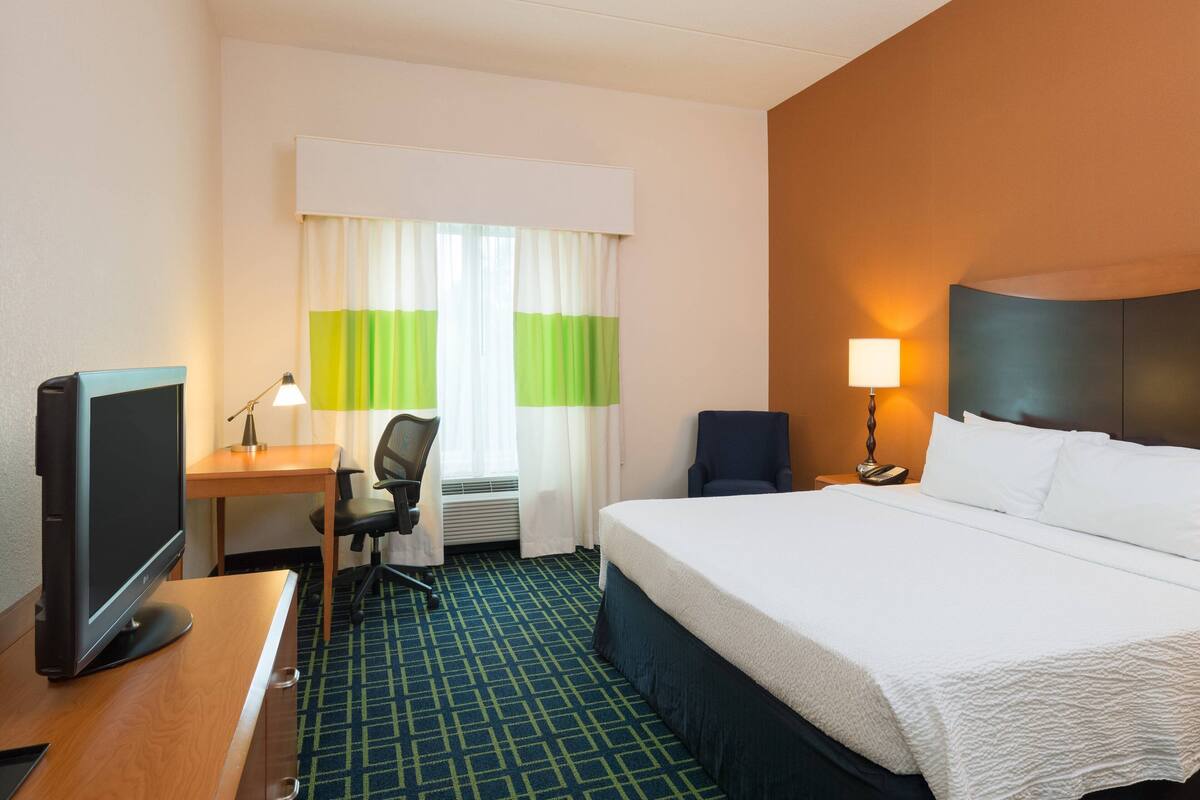 Una habitación de hotel con cama, escritorio, silla, televisión y lámpara.