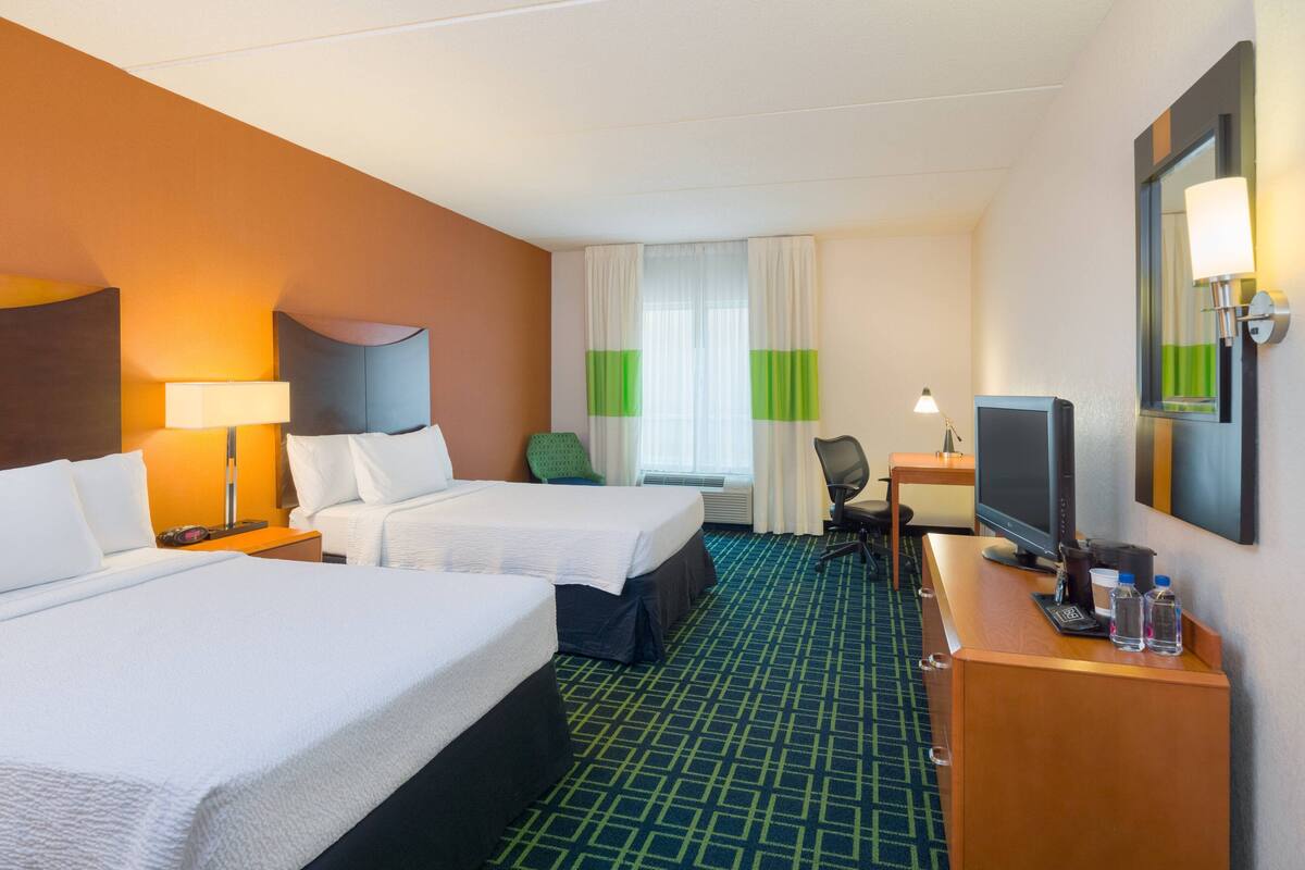 Una habitación de hotel con dos camas, un escritorio con computadora, un televisor y un espejo.