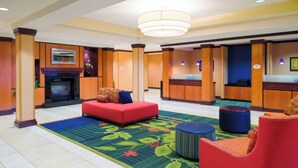 A hotel lobby with a red sofa, a blue ottoman, and a red armchair. There is a reception desk, a fireplace, and a large chandelier.