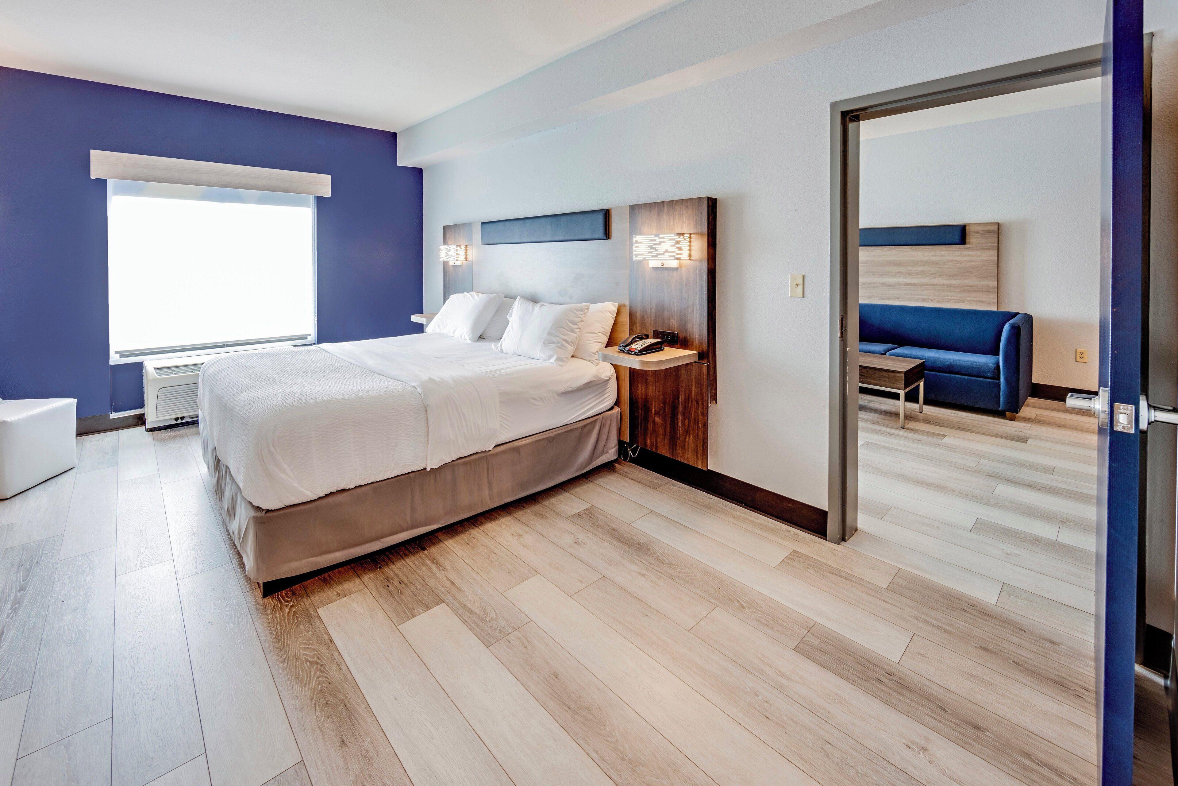 Quarto de hotel moderno com cama grande, mesas de cabeceira, um banco embutido e vista para o quarto vizinho.