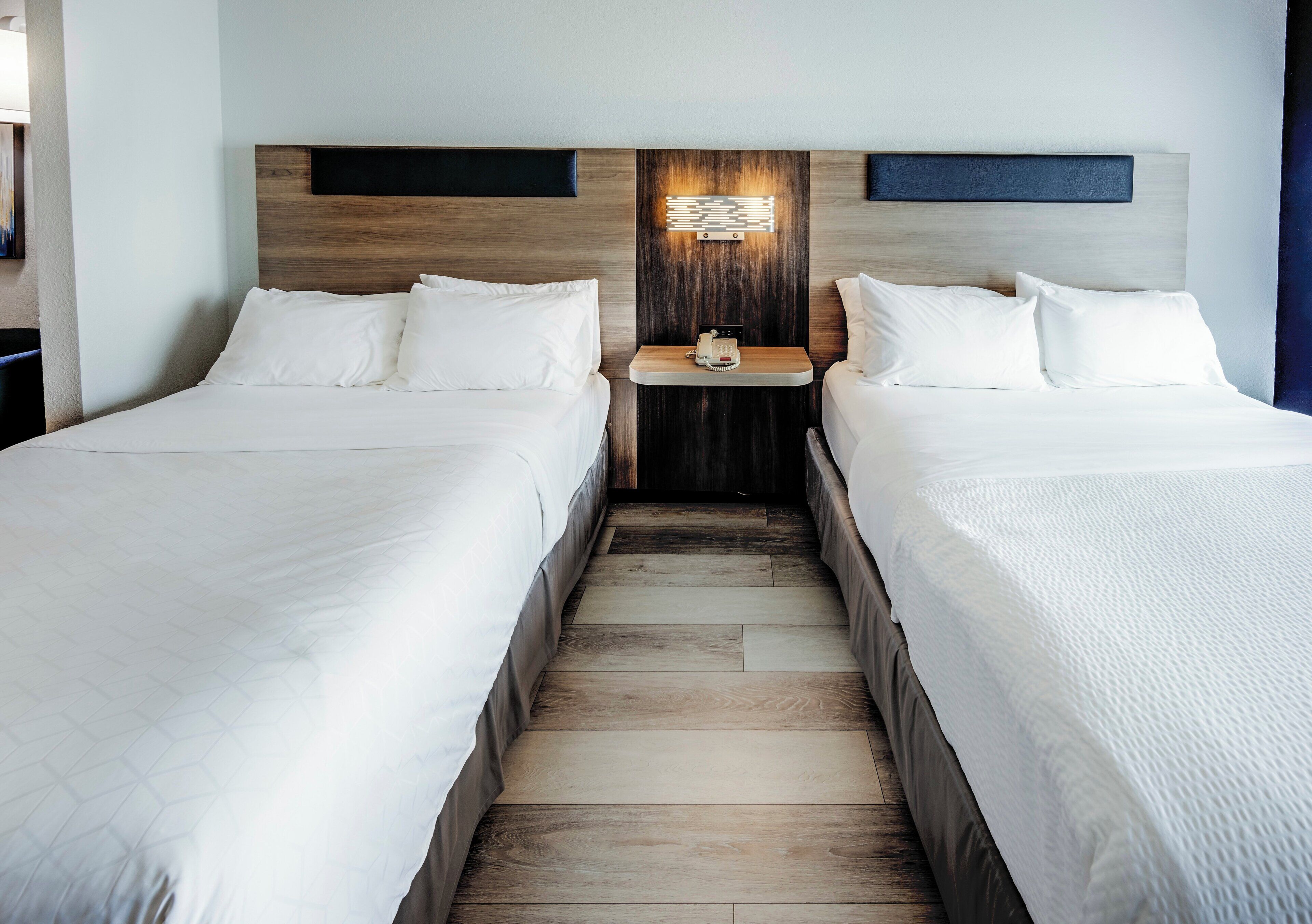 A hotel room with two beds, a wooden headboard, a nightstand with a lamp, and a small table.