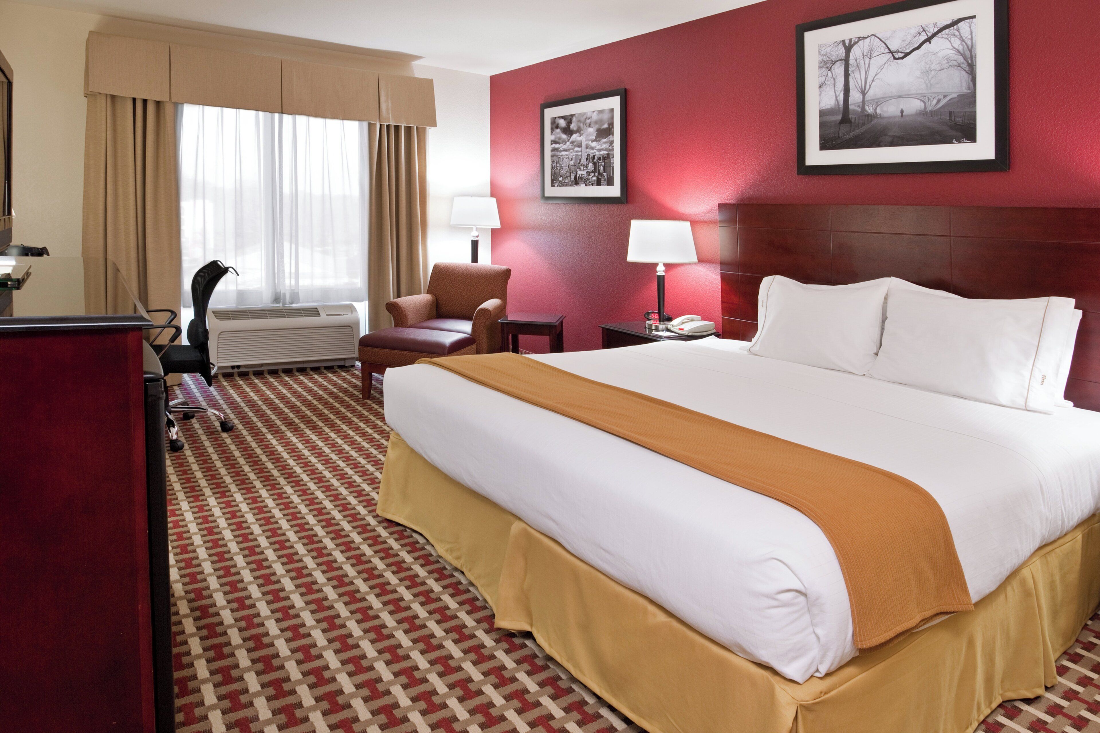 A hotel room with a large bed, a desk, a chair, a lamp, and two framed pictures on the wall.