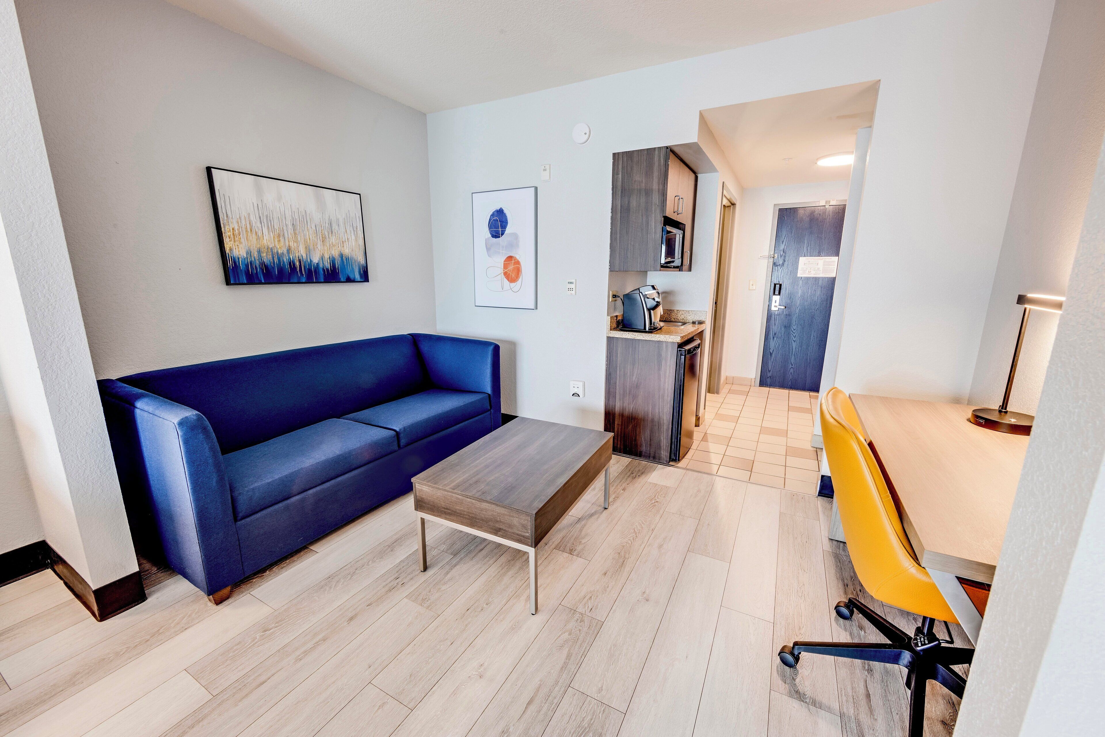 A modern living room with a blue sofa, a wooden coffee table, and a yellow office chair. A kitchen is visible in the background.