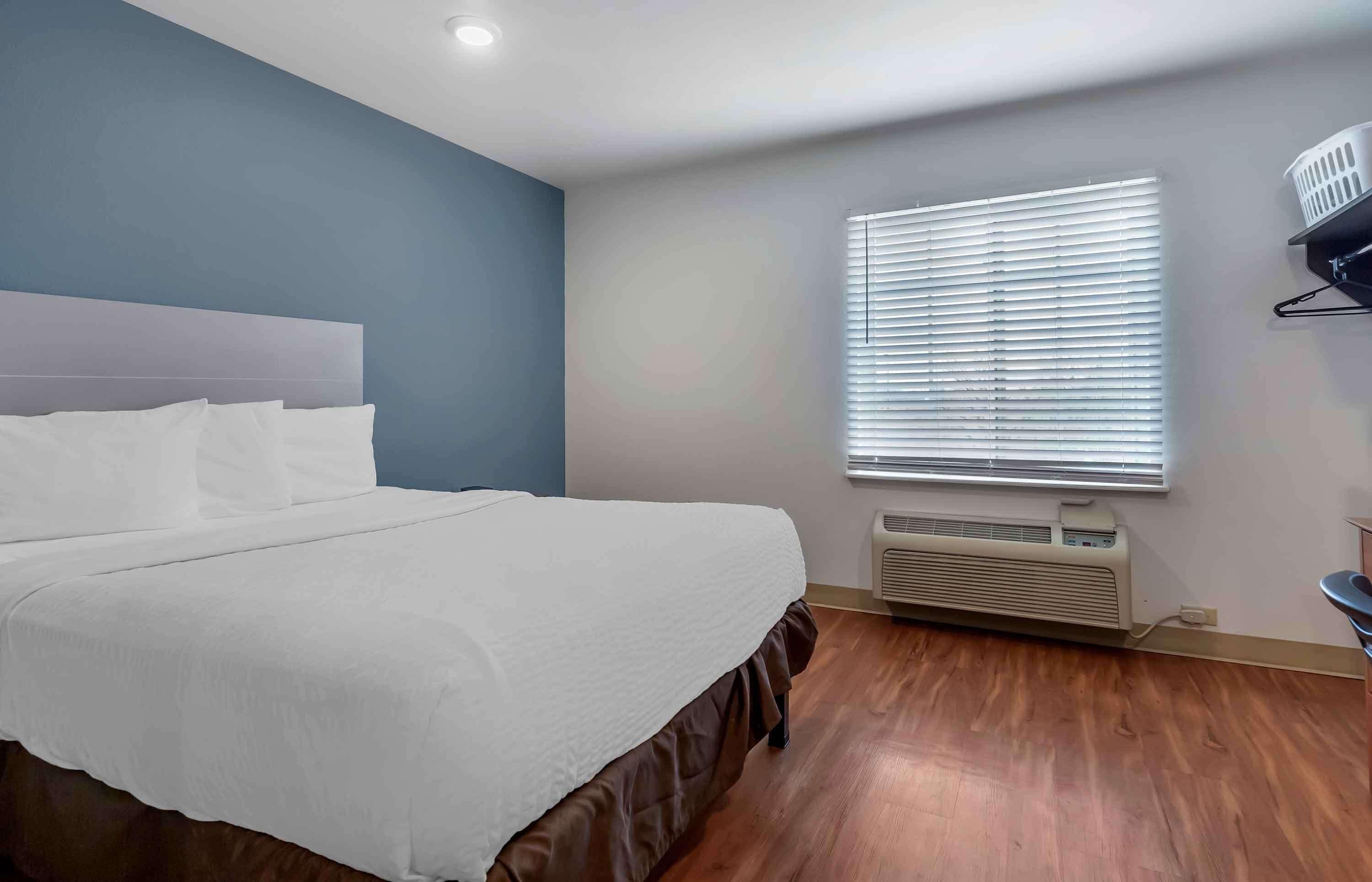 Une chambre d’hôtel avec un grand lit, une fenêtre avec des stores, un climatiseur fixé au mur et un panier à linge accroché au mur.