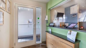 Un área de recepción con puerta de vidrio, un mostrador de recepción con computadora y una pared verde.
