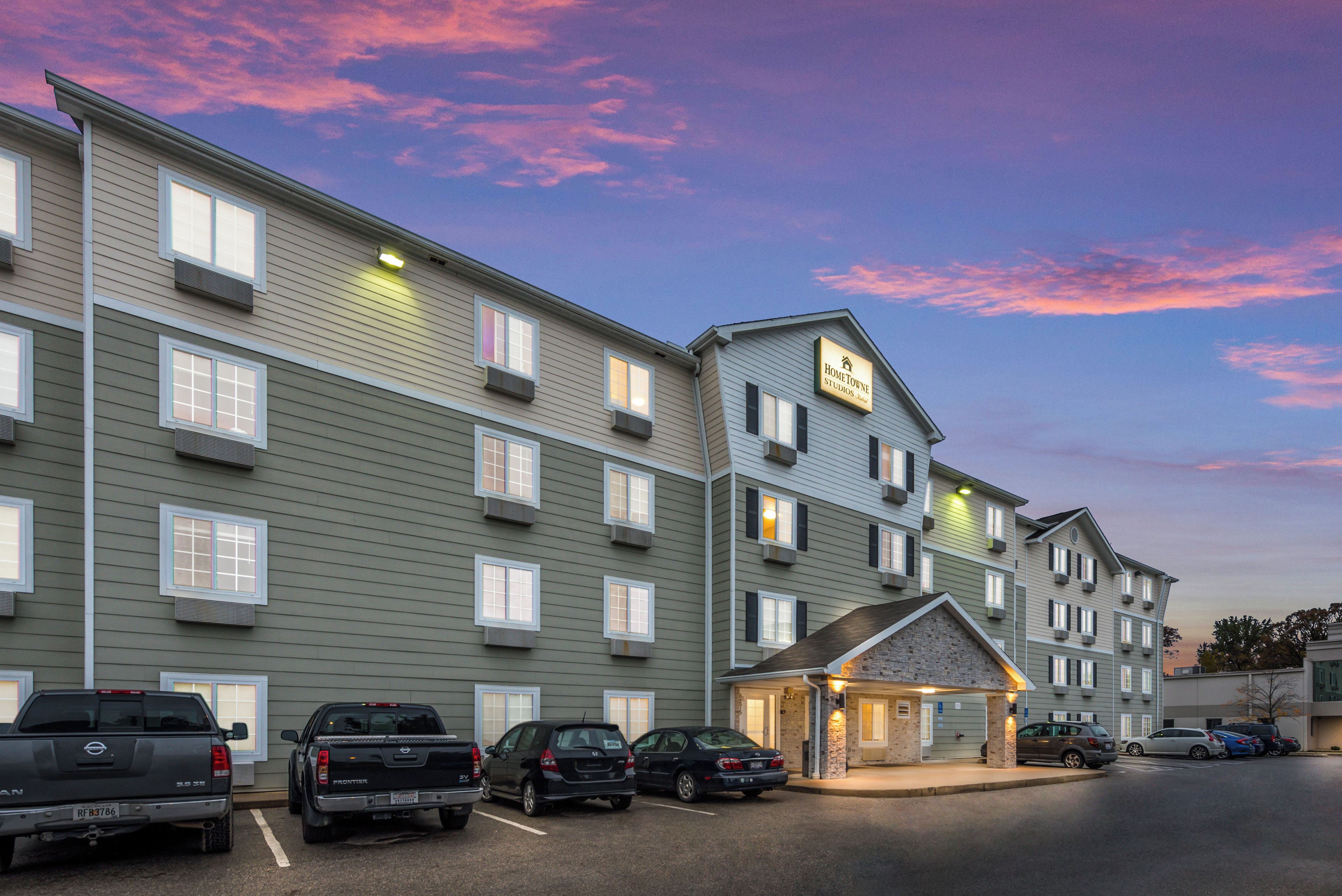 A multi-story hotel with a sign indicating 'HomeTown Studios Hotel.' There are several parked vehicles in front, including a pickup truck and a sedan.