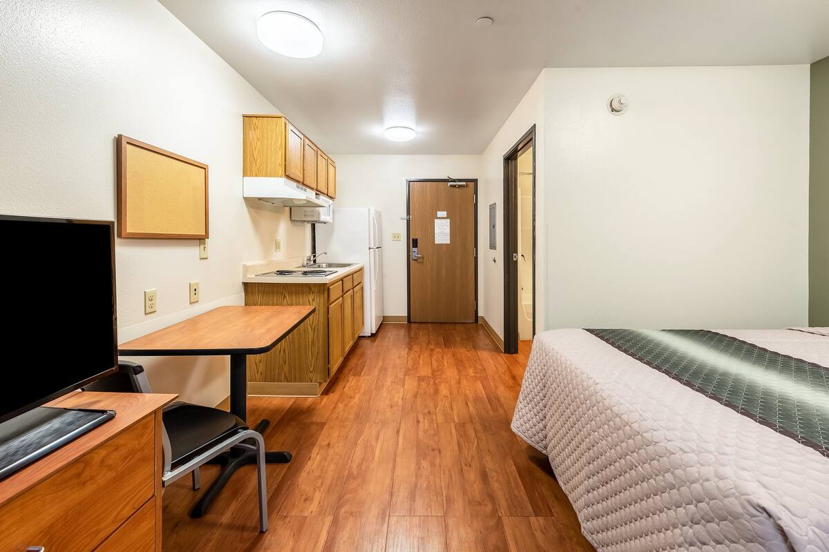A compact hotel room with a kitchenette, a bed with a quilted cover, a desk with a computer, and a television.