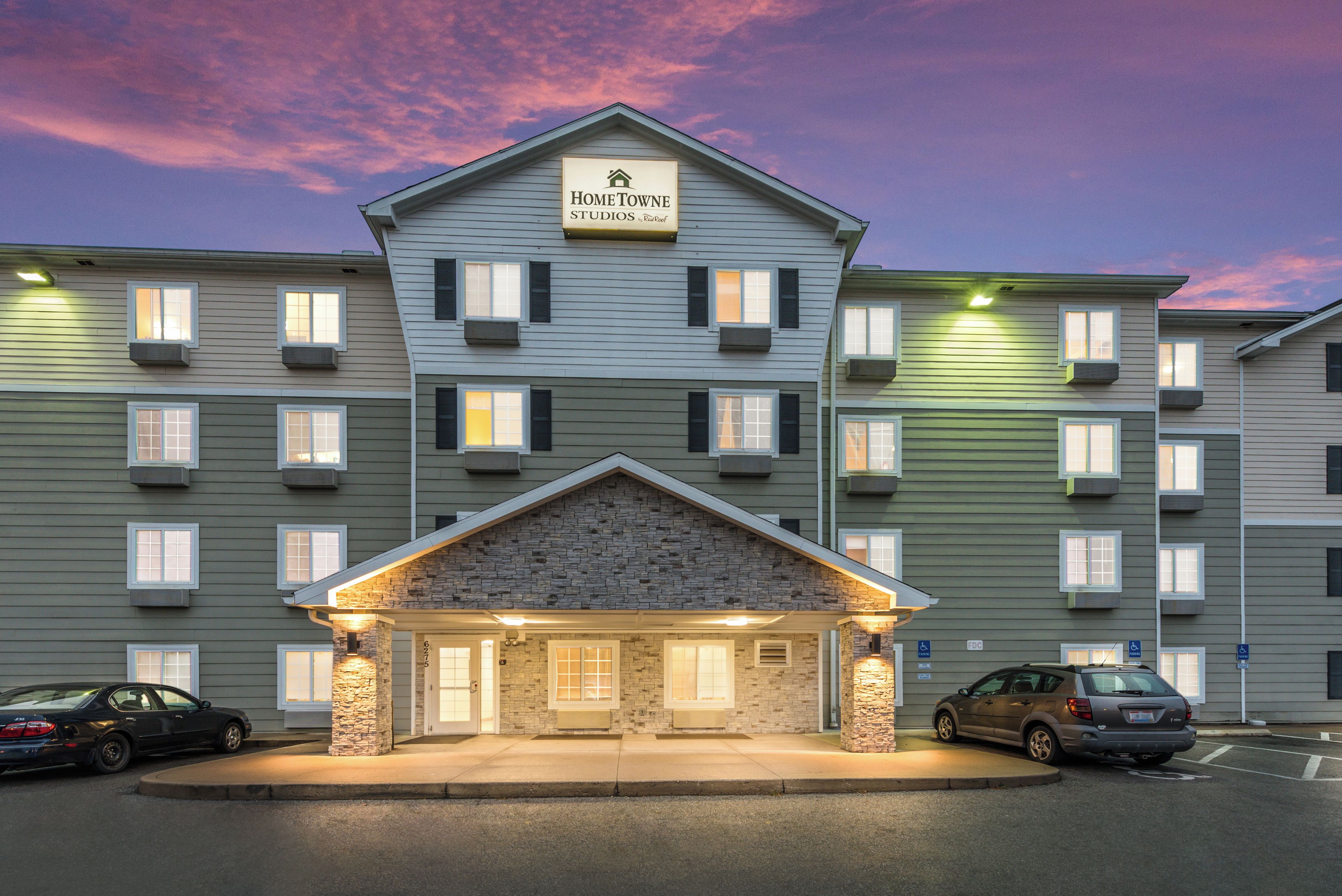 A multi-story hotel with a stone facade entrance, multiple windows, and a sign indicating it is a Hometown Studios by Real Roof property.