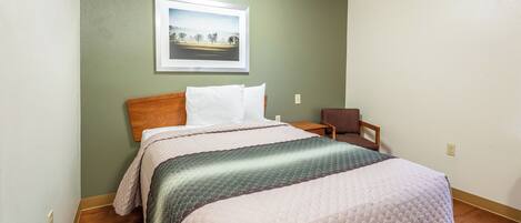 A neatly made bed with a quilt, a framed picture on the wall, and a chair in a room with wooden flooring.