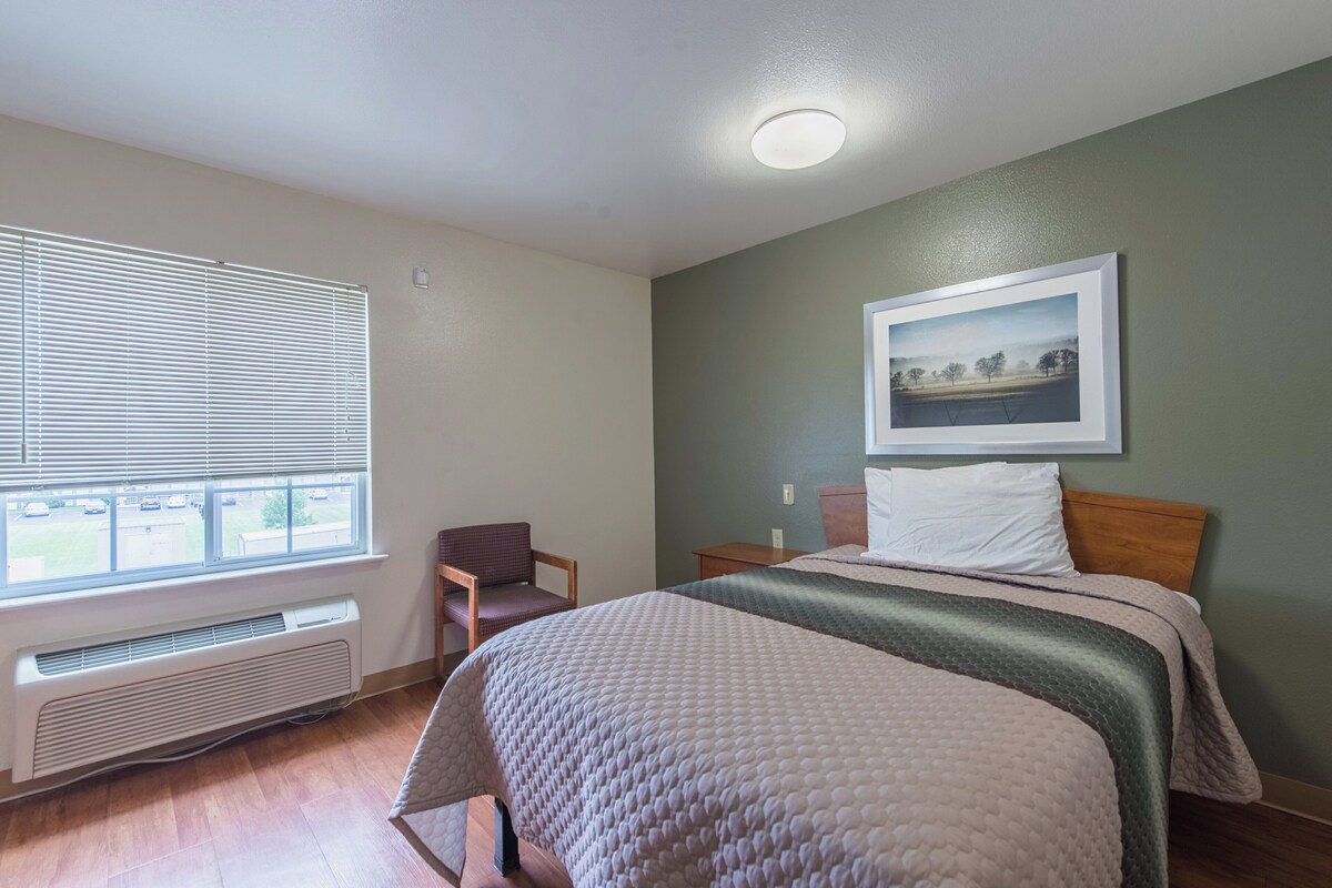 A hotel room with a bed, a chair, a window with blinds, a radiator, and a framed picture on the wall.